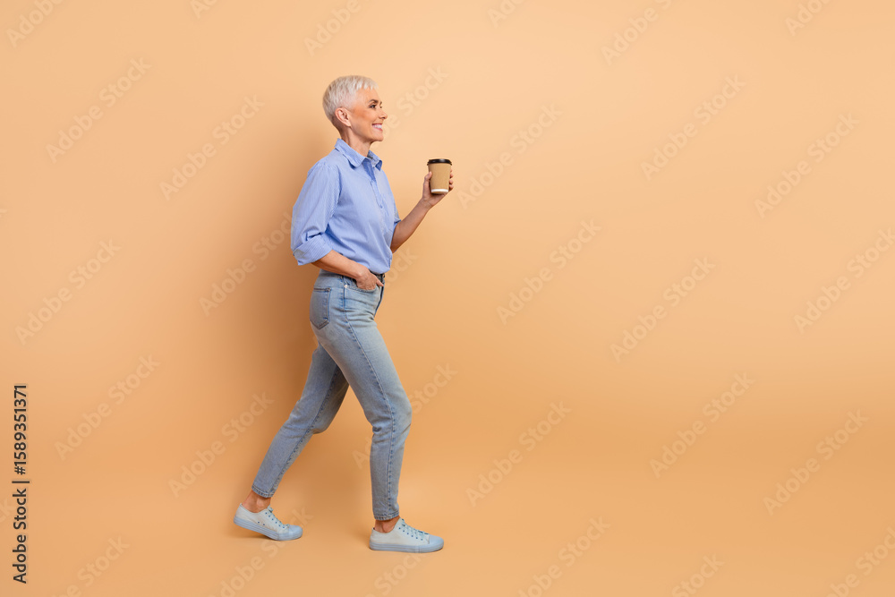 Stylish senior woman walking confidently in casual outfit against beige background while holding a takeaway coffee cup Stylish senior woman walking confidently in casual outfit against beige background while holding a takeaway coffee cup