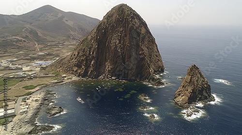 Single isolated rock rising from tranquil blue ocean, drone shot with soft reflections on water, expansive clear sky framing the scene midday