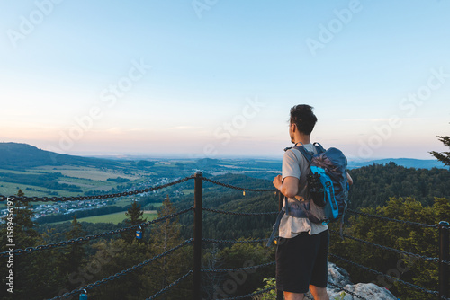 Backpacker stands on a platform at golden hour, overlooking the forested hills and valleys of the Jeseniky Mountains bathed in warm sunset light.