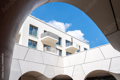 Modern Architecture In Freiham Munich Bavaria: Minimalist Building With Geometric Facade, Large Windows, Curved Arch Details, And Small Tree In Courtyard Under Cloudy Sky