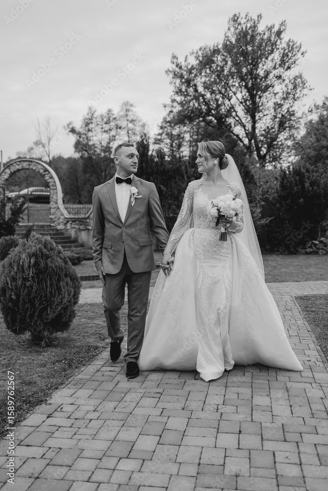 Couple walks hand in hand through garden path on their wedding day under cloudy sky Couple walks hand in hand through garden path on their wedding day under cloudy sky