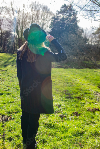 Woman in dark coat and hat walking through sunny meadow with visible lens flare