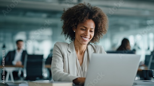 Smiling young African American businesswoman working on a laptop at her desk in a bright modern office with colleagues in the background, no logos, no brands