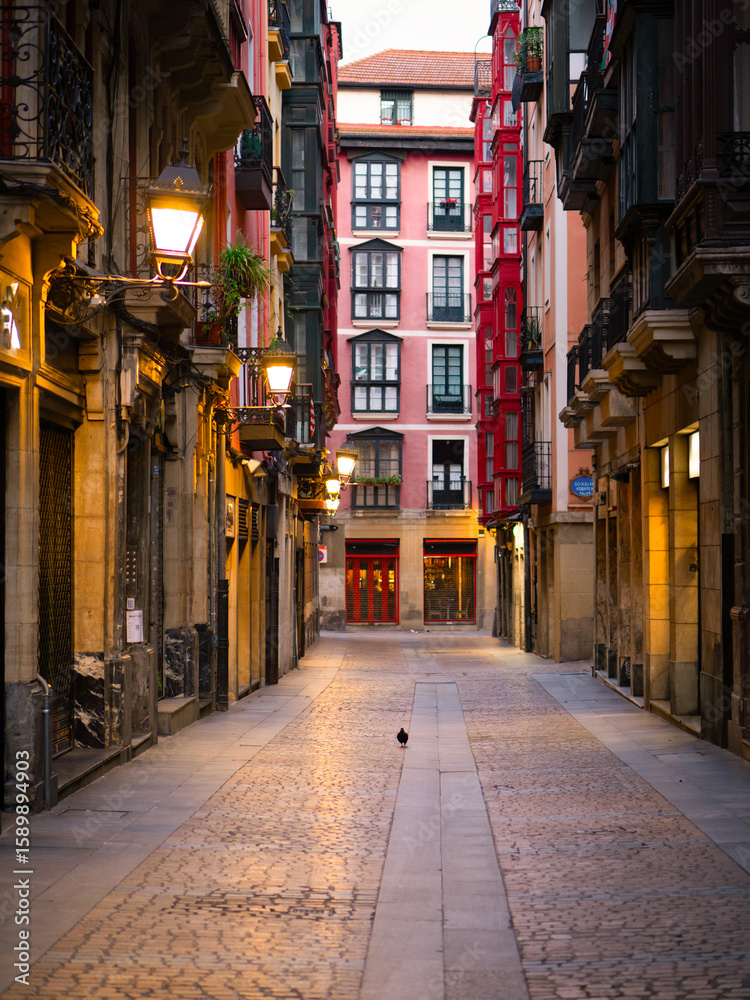 Obraz premium Charming Kapelagile Kalea in Casco Viejo, Bilbao, Spain, with cobblestone streets, historic buildings, and glowing streetlights—perfect for cultural travel and urban exploration.
