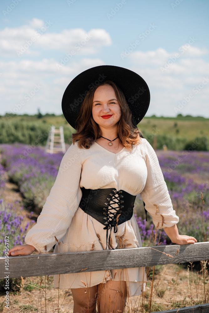 Obraz premium Woman in a white dress and black hat poses in a lavender field on a sunny day during summer