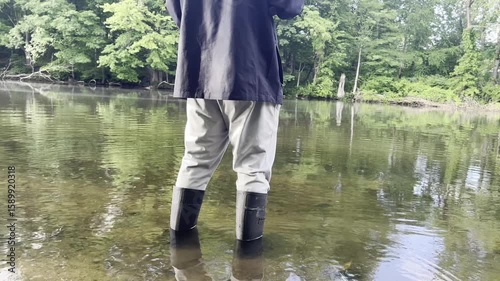 Man stands in shallow lake water wearing rubber boots, holding a fishing rod while casting. Peaceful outdoor fishing scene with calm water and natural surroundings.