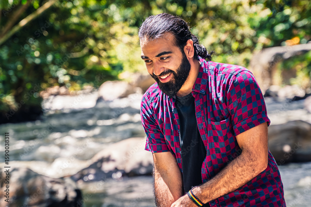 Obraz premium Man in plaid shirt with tied hair standing near river and extending arm toward colorful fence in lush Colombian forest at countryside hotel in Santuario, Risaralda, with space for text