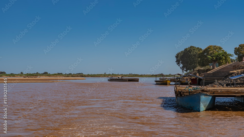 Ferry crossing on an exotic African river. Makeshift berths, ferries made of boats and log decks. Stairs on the hillside. Sandy shores, red-brown water, clear blue sky. Copy space. Madagascar. Ferry crossing on an exotic African river. Makeshift berths, ferries made of boats and log decks. Stairs on the hillside. Sandy shores, red-brown water, clear blue sky. Copy space. Madagascar.