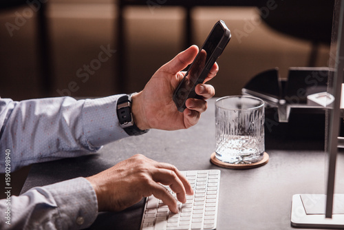 Man using smartphone at modern office desk with keyboard, mouse, monitor, and a glass of water. Minimalist business workspace, perfect for remote work or digital communication.