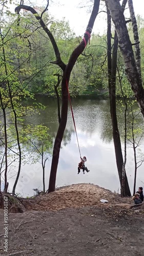 Girls bungee ride over a river in the forest. The rope is tied to a tree, there is a sawdust bank below. Spring landscape, calm water, relaxing in nature.