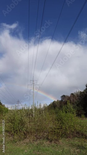 A rainbow in the sky over a green field with bushes and a power line. The contrast of the bright sky, clouds and the rainbow arc creates a lively and dynamic natural and urban scene.