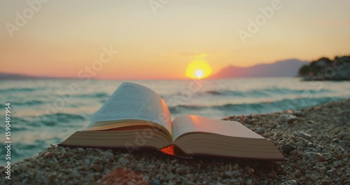 Bible pages turning on pebble beach at sea with glowing sunset light