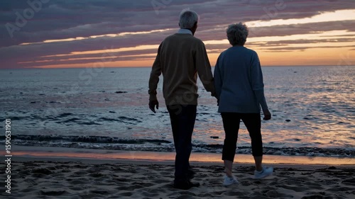 A serene video scene of an elderly couple walking hand in hand on a beach at sunset, captured from a side angle, emphasizing tranquility and companionship.