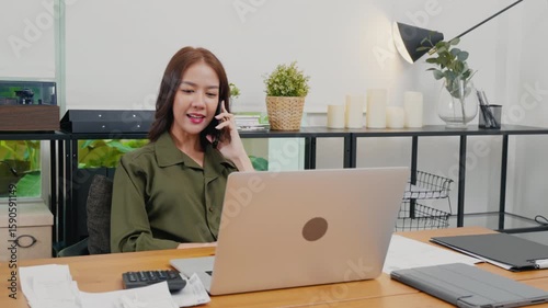 Happy Asian young woman working on laptop computer at home office while talking on mobile phone, smiling businesswoman having business call to talking with company sales client financial