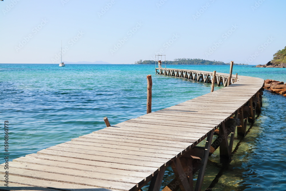 Fototapeta premium Curved wooden pier extending into turquoise waters with a distant yacht and island backdrop in Klong Mad fisherman village, Koh Kood, Thailand.