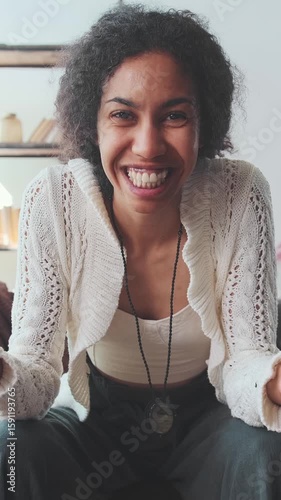 Young beautiful positive African American woman talks with smile and waves hand in greeting looking at screen and recording video interview for potential employer sits on sofa in living room.
