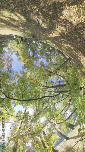 A fisheye lens captures the colorful foliage of trees in a city park during autumn. The video pans across the treetops, revealing a blue sky and puffy white clouds. The leaves are a vibrant mix of