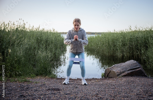 woman doing resistance band squat exercise near lake in summer