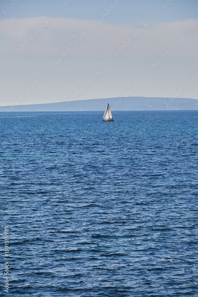 Fototapeta premium White sailboat sails calmly on open blue sea with gentle ripples, distant hills under a soft sky add depth and serenity; sailboat, seascape;