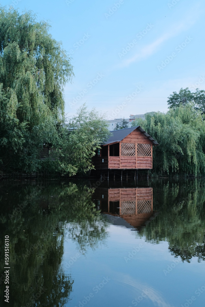 Obraz premium Wooden gazebo on stilts over calm water, surrounded by lush trees, with clear reflections.
