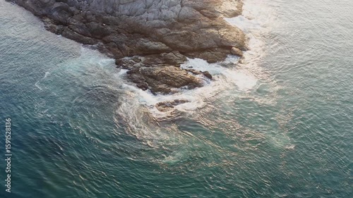 Waves crash powerfully against rugged coastal rocks, showcasing dynamic ocean currents and the raw beauty of nature from above.