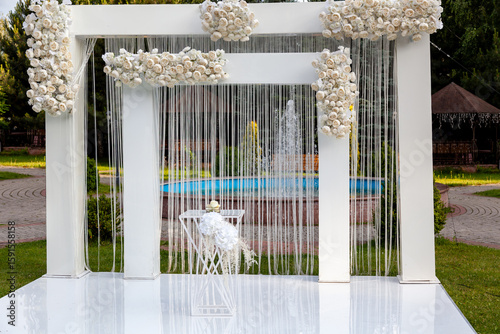 Outdoor wedding setup featuring a modern white arch adorned with cascading white roses and ribbons, positioned in front of a fountain in a scenic garden setting