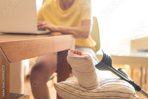 Woman with plastered leg is working on laptop while sitting at home with crutches resting on a pillow, highlighting challenges and adaptation to injury in daily life