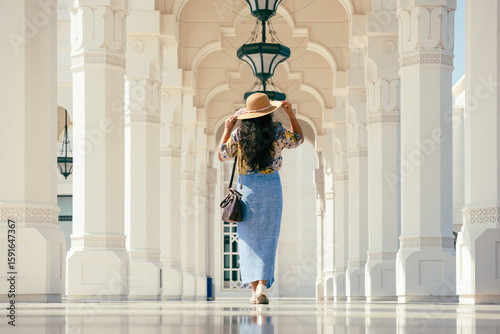 A woman walking under stone arches, wearing a floral blouse, blue skirt, and wide-brimmed hat, carrying a handbag, woman posing for photos 