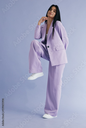 Stylish woman in lavender pantsuit posing confidently against a soft purple background, showcasing contemporary fashion and casual white sneakers in a studio setting.