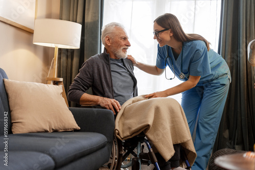 Obraz na plátne Smiling, beautiful nurse covering elderly man in wheelchair with blanket at home