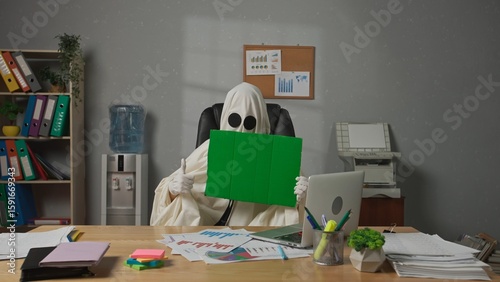 Obraz na plátně Halloween businessman ghost in white sheet sits at office desk holding showing chroma key green screen board at camera