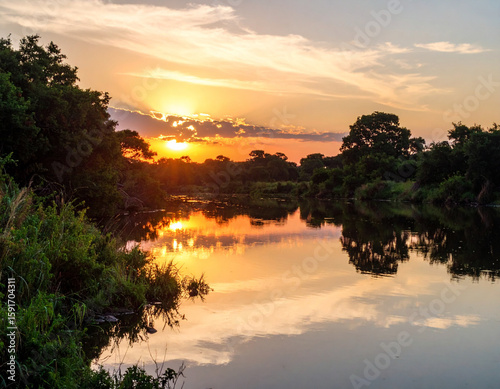 The serene, calm waters of the Kwanza River, reflecting a brilliant sunset sky, framed by lush riverine vegetation.