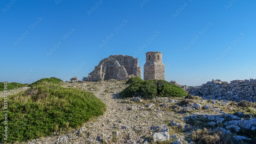 Fototapeta premium Ruins of an ancient stone church with a preserved arch and bell tower on a rocky hill in Kornati National Park, Croatia, under a bright blue sky.