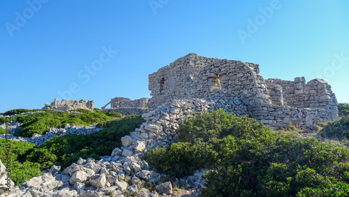 Ancient stone ruins on a rocky hilltop covered with Mediterranean shrubs, under a clear blue sky in Kornati National Park, Croatia.