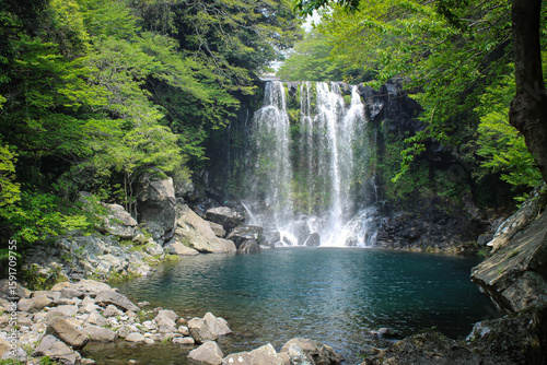Front view of Cheonjeyeon Waterfall cascading into a clear blue pool, surrounded by lush green forest and rocks, Jeju Island, South Korea.