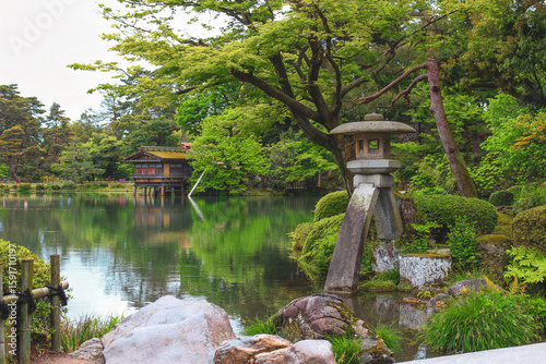 Traditional Japanese stone lantern by a pond in Kenrokuen Garden, Kanazawa, with lush greenery and a wooden teahouse in the background.