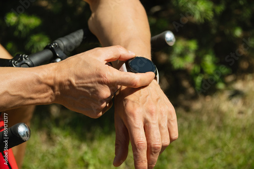 Athlete cyclist checking distance, pulse or fitness progress in apps after riding. Close up man hands checking his heartbeat with smartwatch. Summer activity.