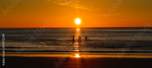 Three people enjoying time in the seawater silhouetted against vibrant midnight sun. Lofoten Islands, Northern Norway