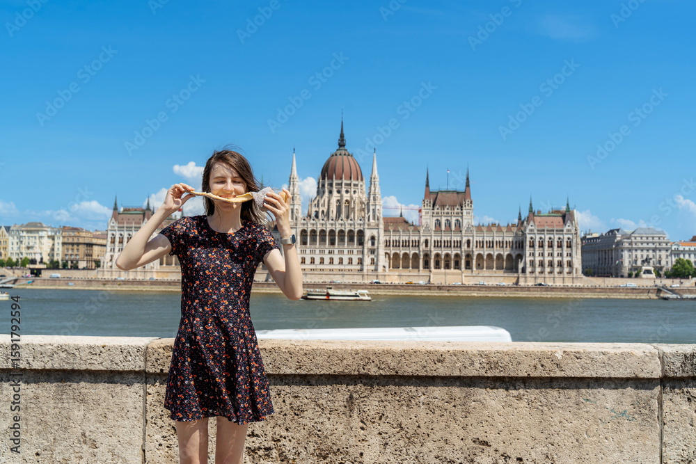 Obraz premium Woman Enjoying Kürtőskalács in Front of Budapest Parliament