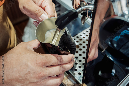 Close-up of barista steaming milk using a metal frothing pitcher and espresso machine steam wand for a hot coffee drink.