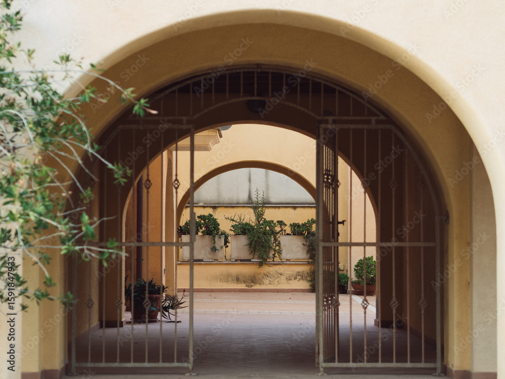 Fototapeta premium Archway with Potted Plants and Metal Fence – Italian Apartment Building in Golfo Aranci, Sardinia, Horizontal Photo