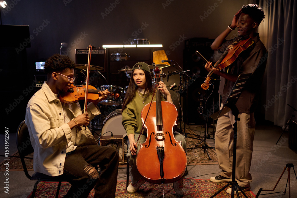 Fototapeta premium Multiethnic group of young musicians playing string instruments in music studio, Black boy playing violin, Hispanic girl holding cello, Black man standing with violin