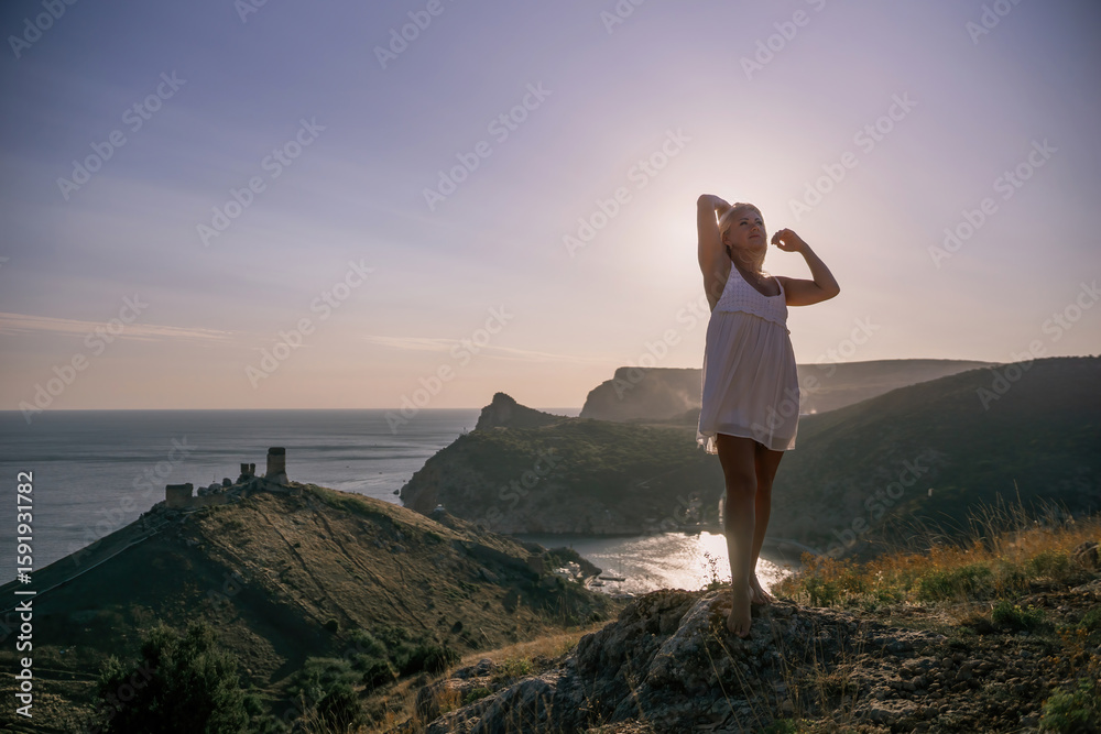 Fototapeta premium A blonde woman stands on a hill overlooking the ocean. She is wearing a white dress and she is enjoying the view.