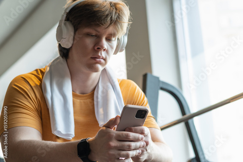 Sporty young man relax and sitting with mobile phone, listen music headphones and texting at gym. Bodybuilder during training . Fitness lifestyle concept. Take a break, looking at camera.