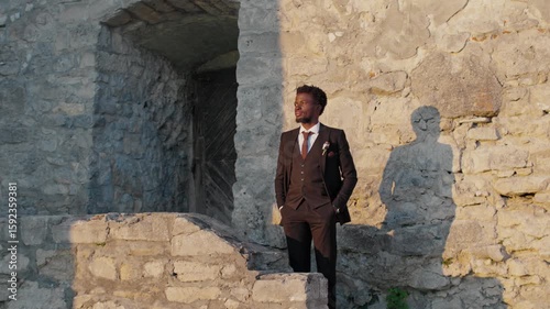 Portrait of African American groom standing in sunlight near rustic stone wall, hands in pockets, casting strong shadow. Groom posing confidently alone before wedding ceremony at old castle ruins.