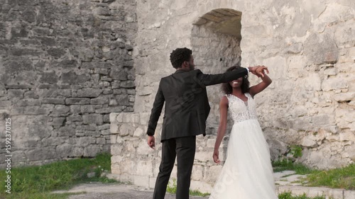 Bride and groom holding hands while walking, groom glancing back smiling. African American couple enjoying romantic moment near ancient stone wall during wedding photo session outdoors.