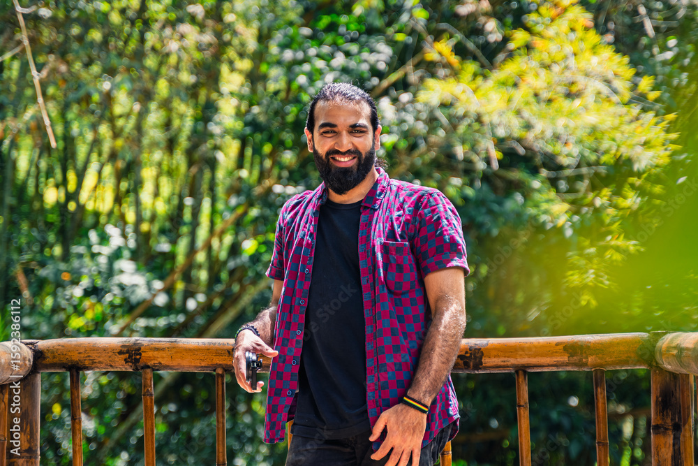 Obraz premium Colombian man with tied hair looking at camera while holding camera on wooden railing in lush forest at countryside hotel in Santuario, Risaralda, with space for text