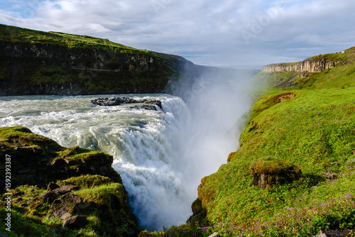 Water flows powerfully over the Gullfoss waterfall in Iceland, creating mist that dances with sunlight.