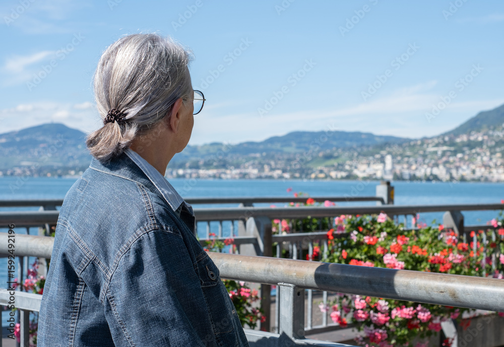 Fototapeta premium Rear view of a gray-haired senior woman sitting in front of the Geneva lake admiring the landscape, a relaxed senior grandmother enjoying peaceful moments in retirement