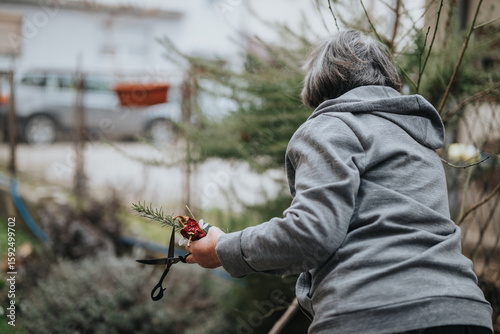 An elderly woman wearing a hoodie tends to her garden, using pruning shears to clip branches while holding flowers. The outdoor setting is calm and serene, featuring a natural environment.